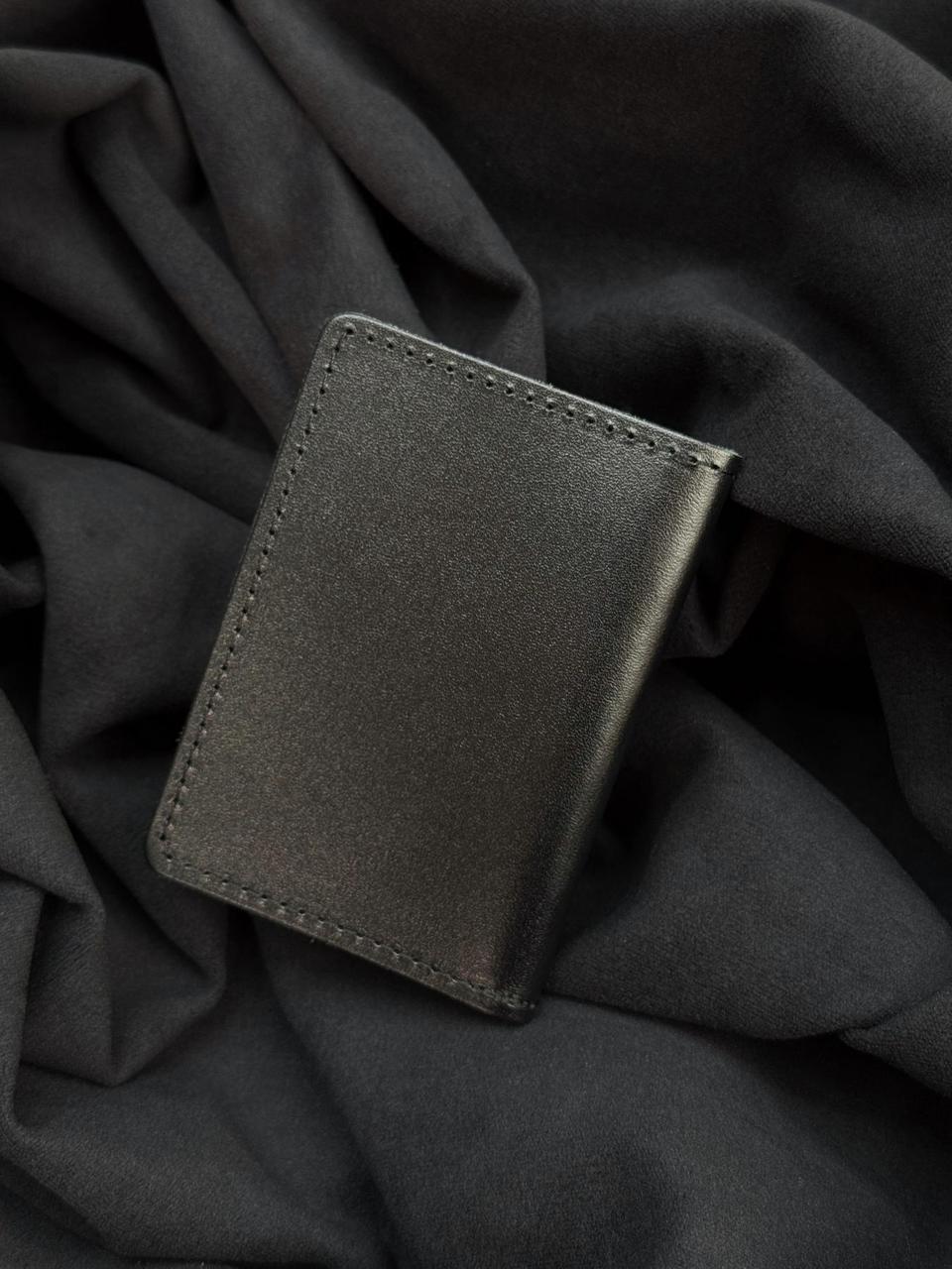 A slim black leather card holder or wallet with visible stitching, resting on a dark gray draped fabric background.