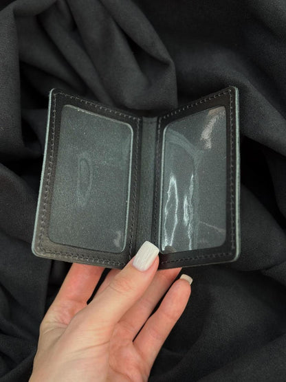 A black leather bi-fold wallet with two clear plastic ID slots, held open by a hand against a dark gray fabric background.