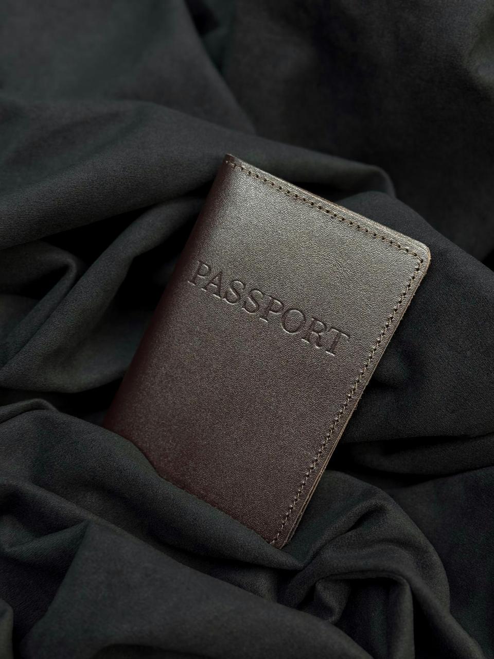 A dark brown leather passport holder with 'PASSPORT' embossed on the front, resting on draped black fabric.