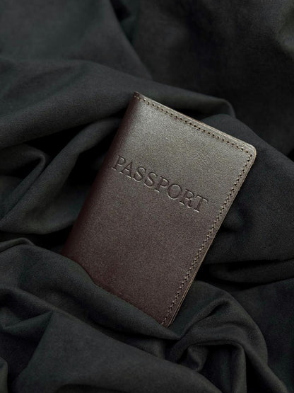 A dark brown leather passport holder with 'PASSPORT' embossed on the front, resting on draped black fabric.