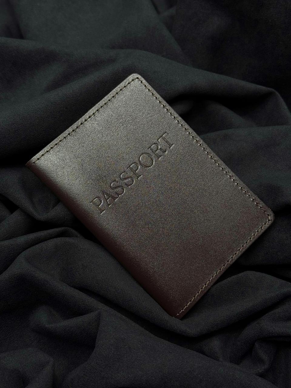 A dark brown leather passport holder with 'PASSPORT' embossed on the front, resting on a dark draped fabric background.