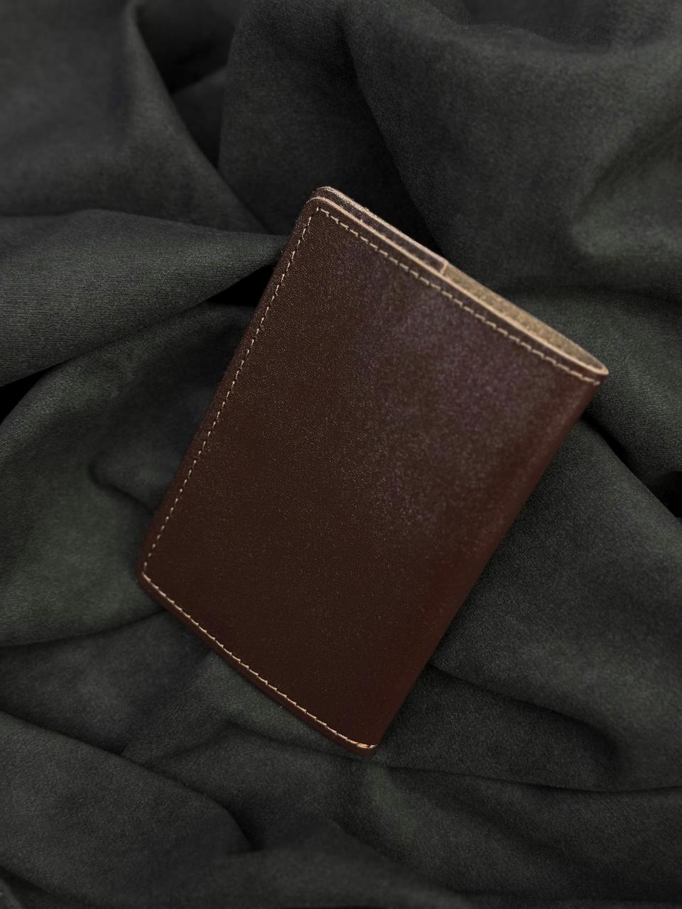 A dark brown leather card holder or wallet with visible stitching, resting on a dark gray draped fabric background.