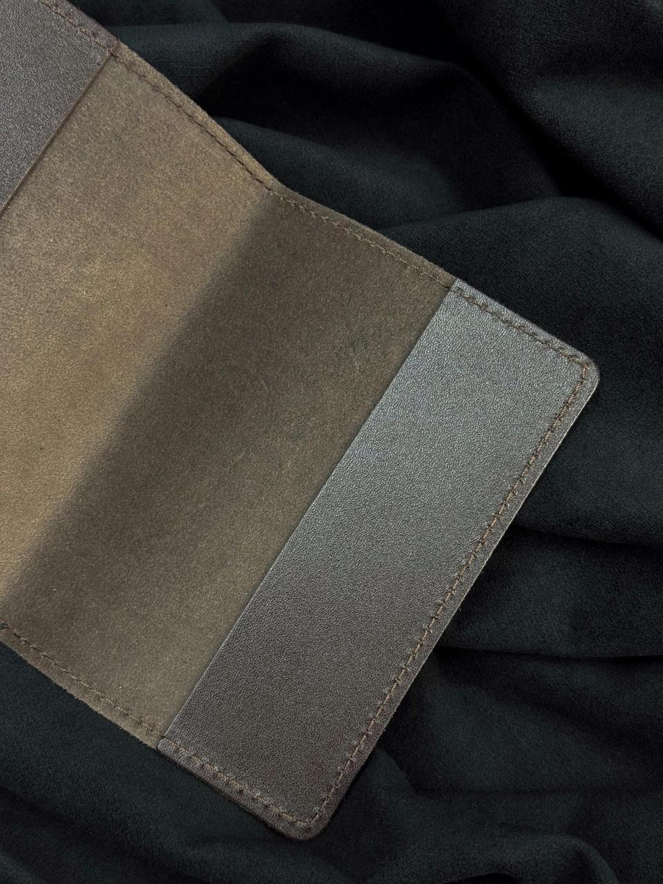 A close-up of a textured leather cardholder or wallet in taupe and gray-brown tones, laid flat on a dark charcoal fabric background.