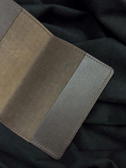 A close-up of a textured leather cardholder or wallet in taupe and gray-brown tones, laid flat on a dark charcoal fabric background.
