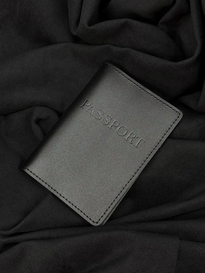 A dark gray faux-leather passport cover with 'PASSPORT' embossed on the front, resting on a draped black fabric background.