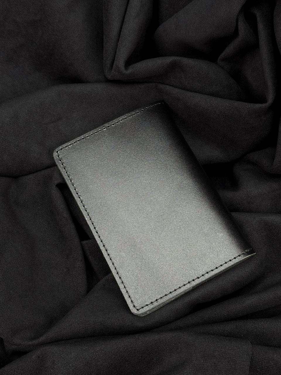 A minimalist black leather card holder wallet with visible stitching, resting on a dark, draped fabric background.
