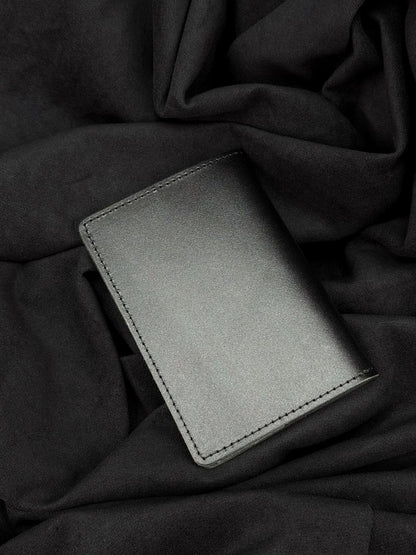 A minimalist black leather card holder wallet with visible stitching, resting on a dark, draped fabric background.