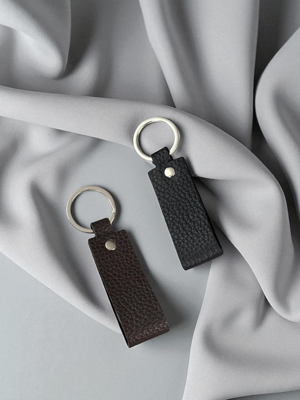 Two pebbled leather keychains in dark brown and black, each with a silver-tone split ring and metal rivet, displayed on draped light gray fabric.