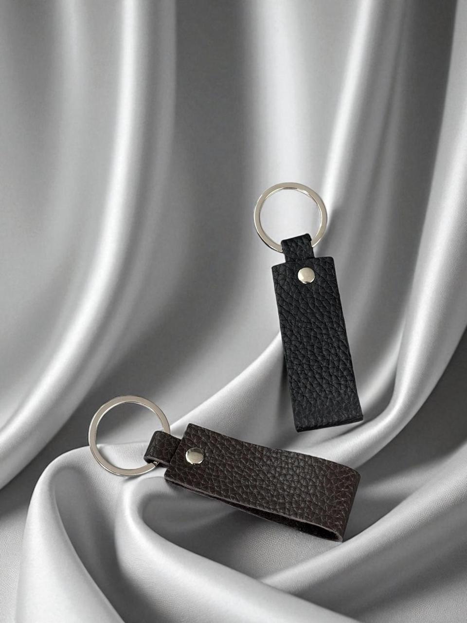 Two leather keychains with silver metal key rings, one in black pebbled leather and one in dark brown pebbled leather, displayed on draped silver satin fabric.