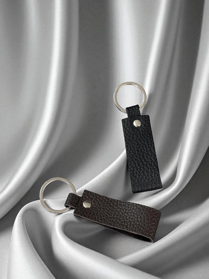Two leather keychains with silver metal key rings, one in black pebbled leather and one in dark brown pebbled leather, displayed on draped silver satin fabric.