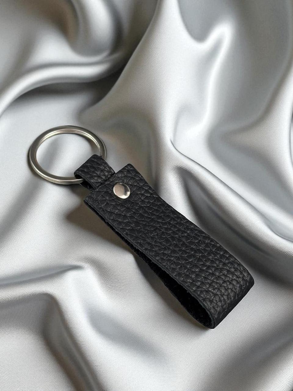 A black textured leather keychain with a silver metal split ring and snap button, displayed on a draped silver satin fabric background.