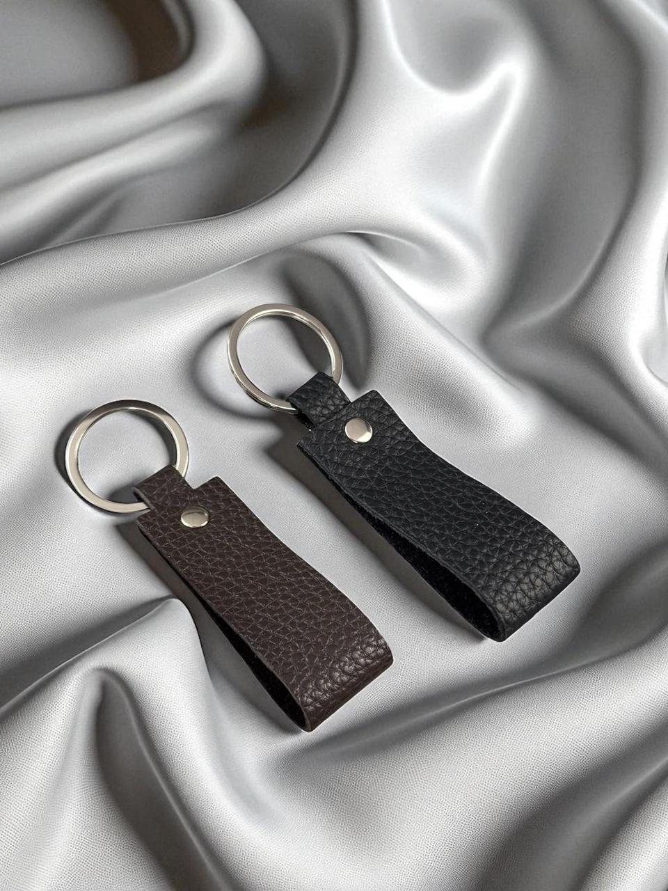 Two pebbled leather keychains, one dark brown and one black, with silver metal key rings, displayed on a draped silver satin fabric background.