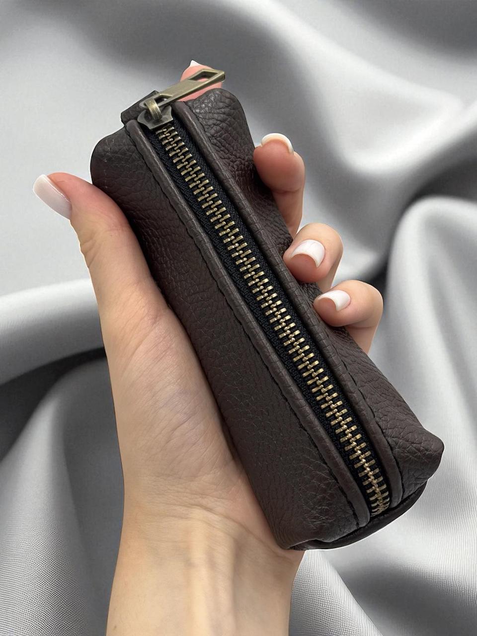 A hand holding a dark brown textured leather zippered pouch with a gold-tone zipper, set against a draped gray satin fabric background.