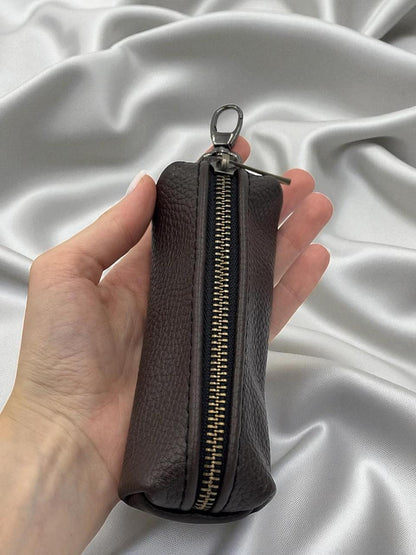 A dark brown textured leather zippered pouch held in a hand against a draped white satin fabric background.