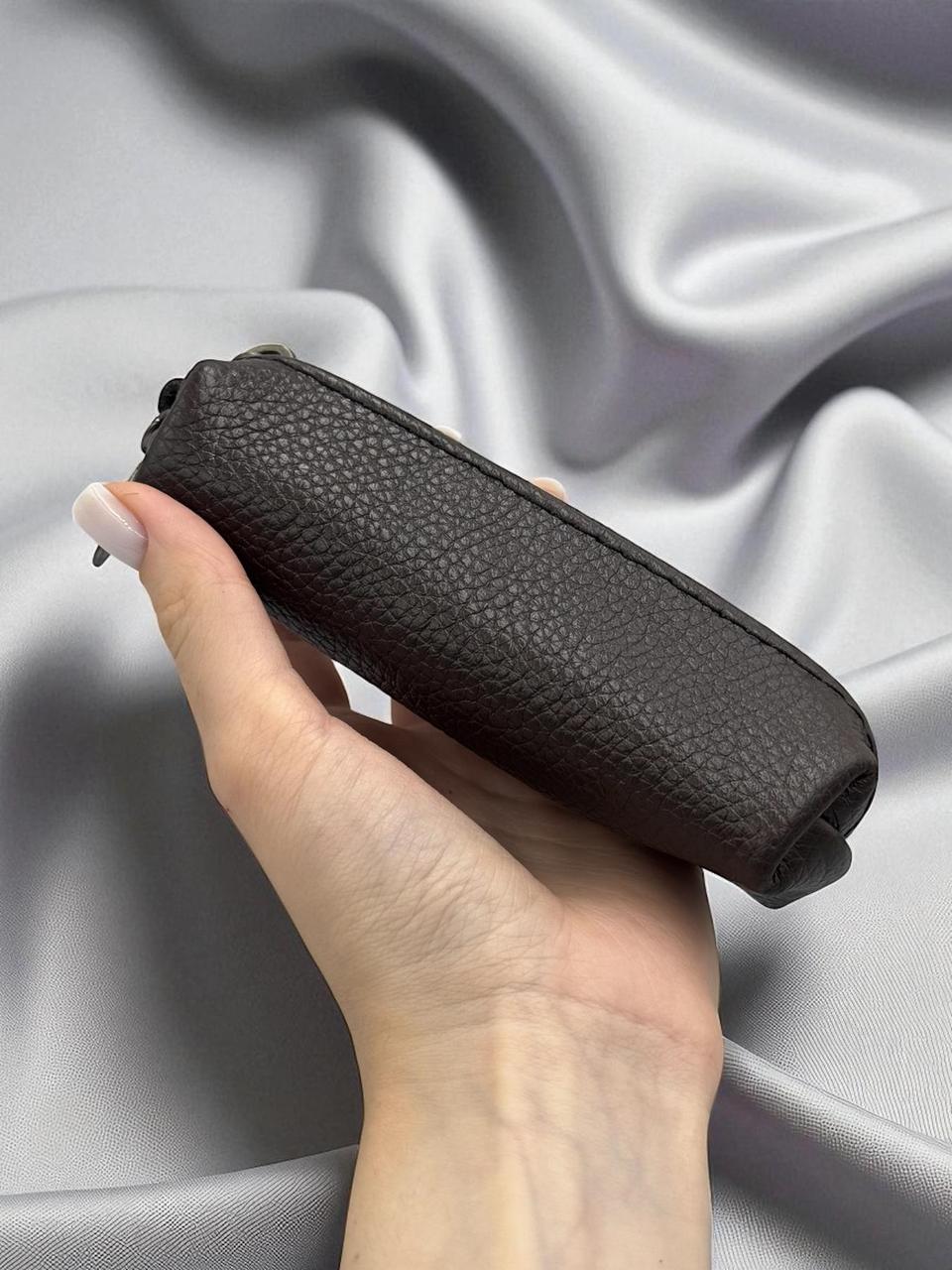 A hand holding a dark brown pebbled leather cylindrical pouch or case against a draped silver satin fabric background.