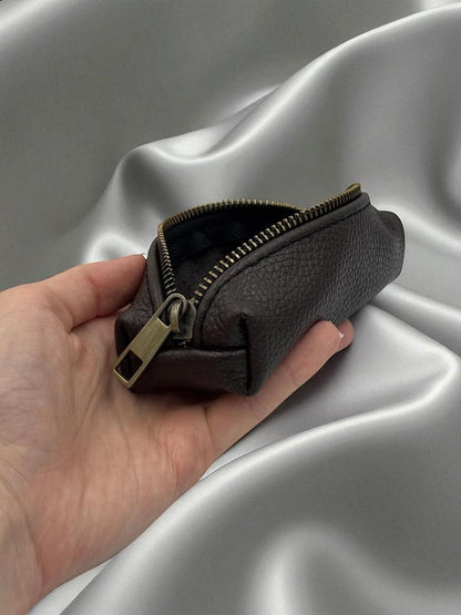 A small pebbled leather zip pouch in dark brown, held in a hand against a draped silver satin fabric background.