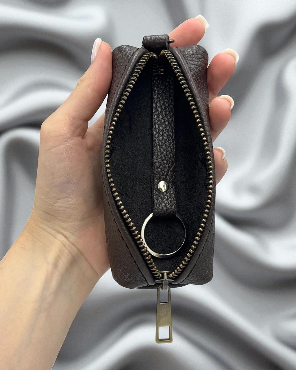 A hand holding an open dark brown pebbled leather zippered pouch with a silver-tone metal keyring, set against a draped gray fabric background.