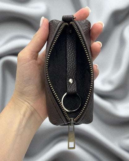 A hand holding an open dark brown pebbled leather zippered pouch with a silver-tone metal keyring, set against a draped gray fabric background.