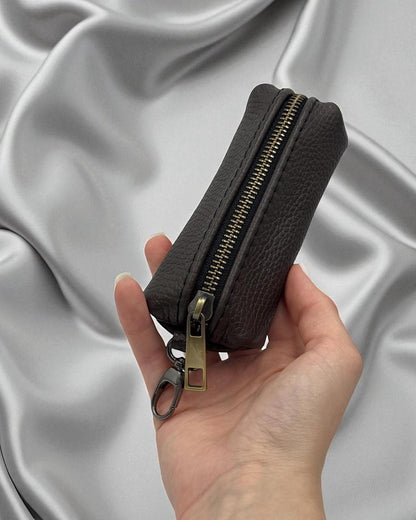 A hand holding a small dark brown pebbled leather zippered pouch with a gold-tone zipper and keychain ring, set against a draped silver satin fabric background.