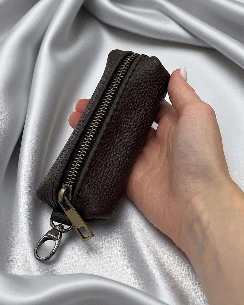 A dark brown pebbled leather zippered pouch with a gold-tone zipper and silver keychain ring, held in a hand against a draped silver satin background.