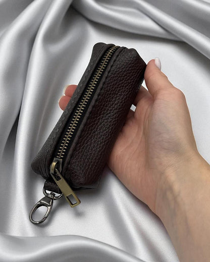 A dark brown pebbled leather zippered pouch with a gold-tone zipper and silver keychain ring, held in a hand against a draped silver satin background.