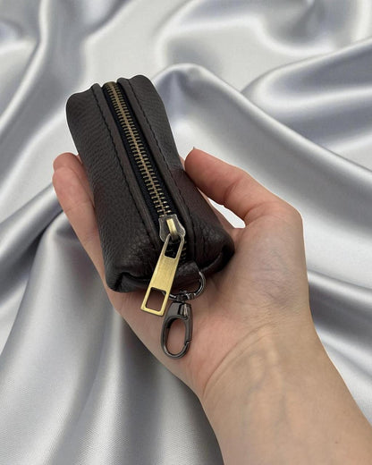 A hand holding a small black leather zip pouch with a gold-tone zipper and keychain clip, set against a draped silver satin fabric background.