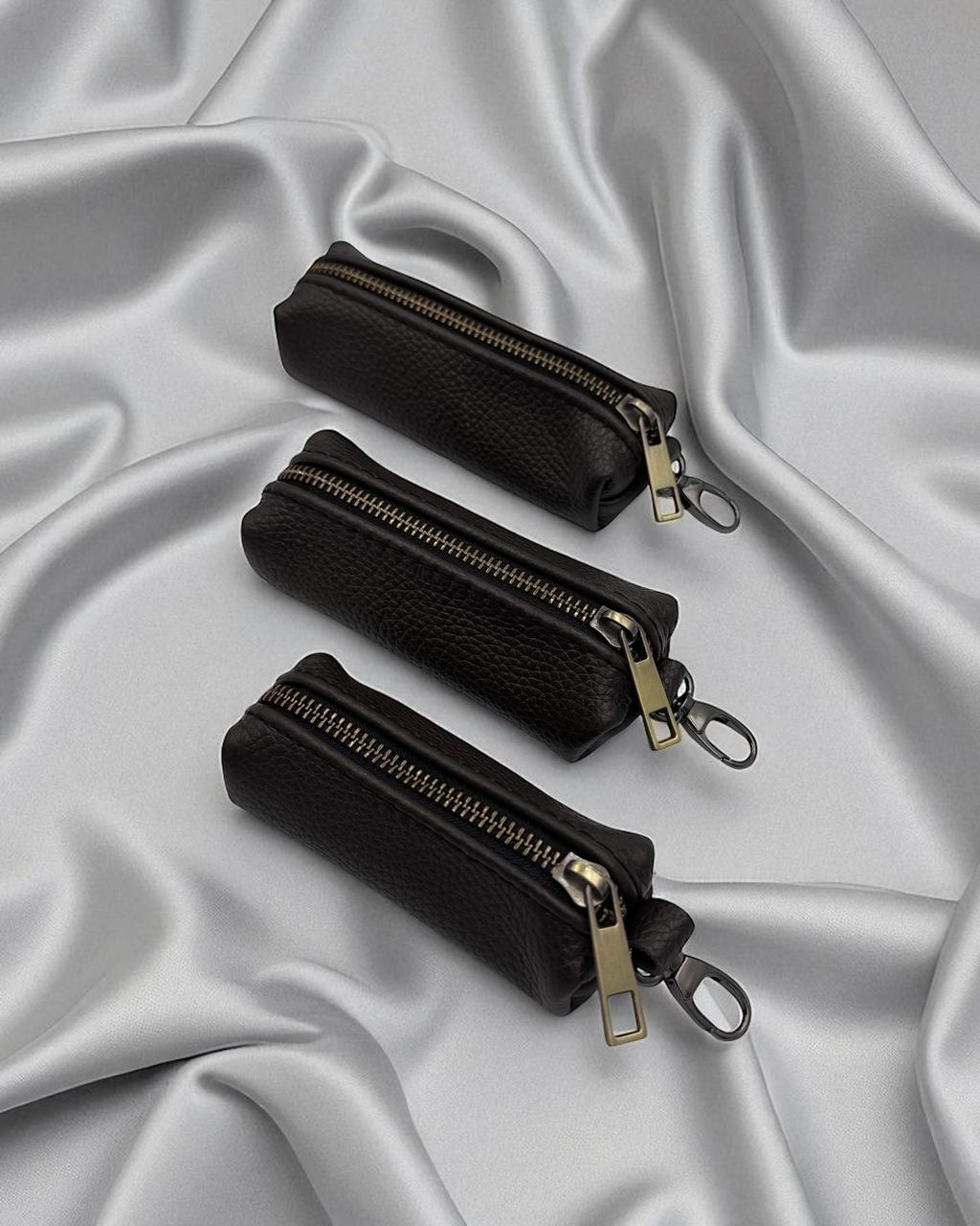 Three small black zippered pouches with gold-tone hardware arranged diagonally on draped silver satin fabric.