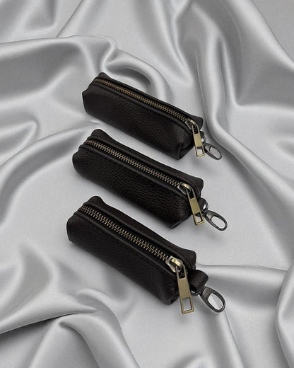 Three small black zippered pouches with gold-tone hardware arranged diagonally on draped silver satin fabric.
