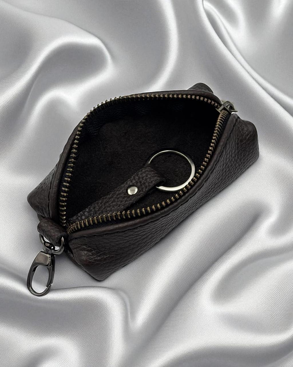 A dark brown textured leather zippered pouch with a key ring attachment, shown open on a draped silver satin fabric background.