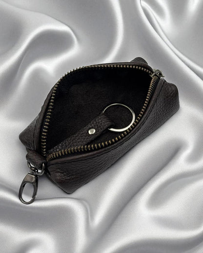 A dark brown textured leather zippered pouch with a key ring attachment, shown open on a draped silver satin fabric background.