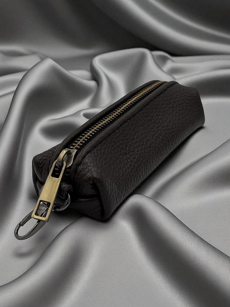 A black pebbled leather zippered pouch with a gold-tone metal zipper pull and key ring, resting on draped gray satin fabric.