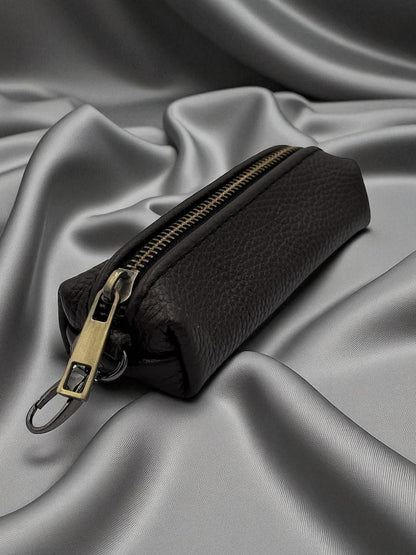 A black pebbled leather zippered pouch with a gold-tone metal zipper pull and key ring, resting on draped gray satin fabric.