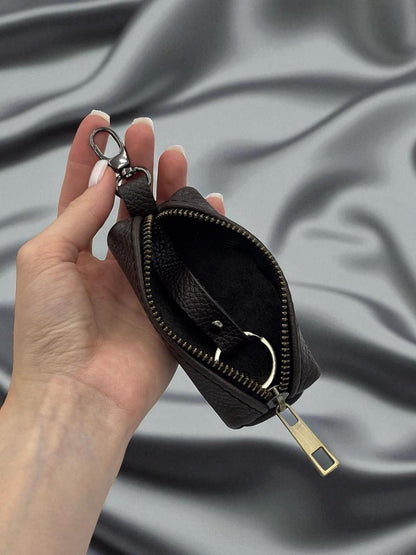A hand holds a small black leather zip pouch with a key ring clasp against a draped gray satin fabric background.