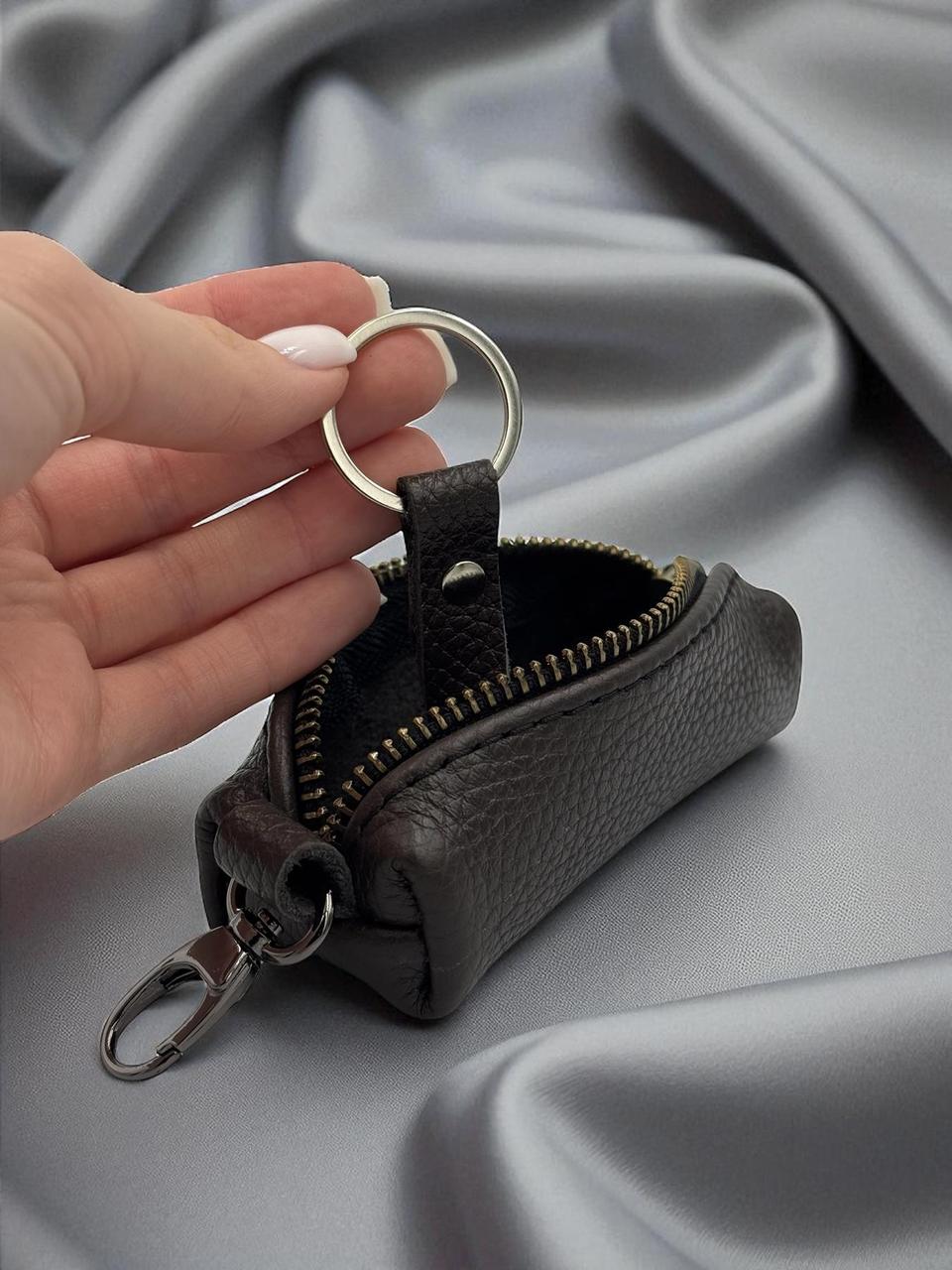 A hand with light pink manicured nails holds a small dark brown pebbled leather key pouch with a silver metal key ring and clip, set against a draped gray satin fabric background.