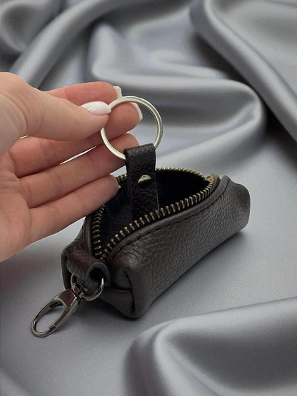 A hand with light pink manicured nails holds a small dark brown pebbled leather key pouch with a silver metal key ring and clip, set against a draped gray satin fabric background.