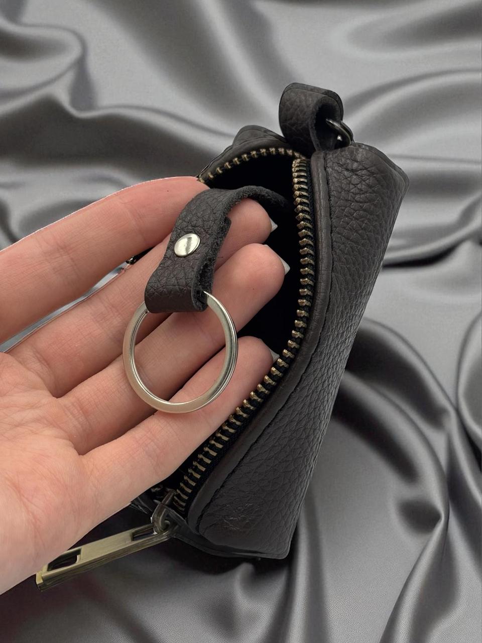 A close-up of a hand holding a small black textured leather zip pouch with a silver metal key ring and zipper pull, set against a draped gray satin background.