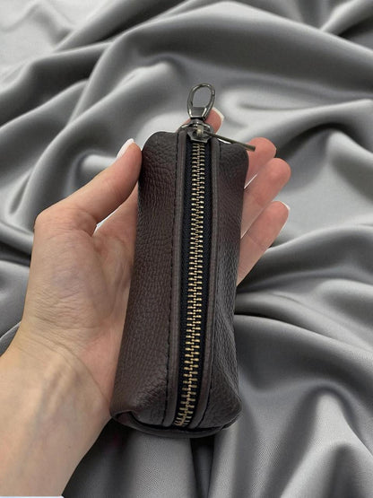 A hand holding a small dark brown pebbled leather zip pouch with a gold-tone zipper and silver keyring clasp, set against a draped gray fabric background.