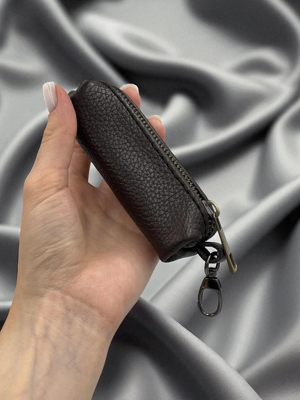 A hand holding a small dark brown pebbled leather zippered pouch with a silver-tone metal clasp, set against a draped gray satin fabric background.