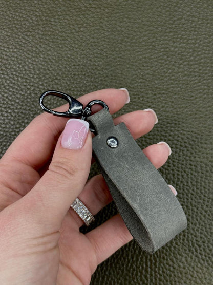 A close-up of a hand holding a gray leather keychain with a black metal lobster clasp against a textured olive green background.