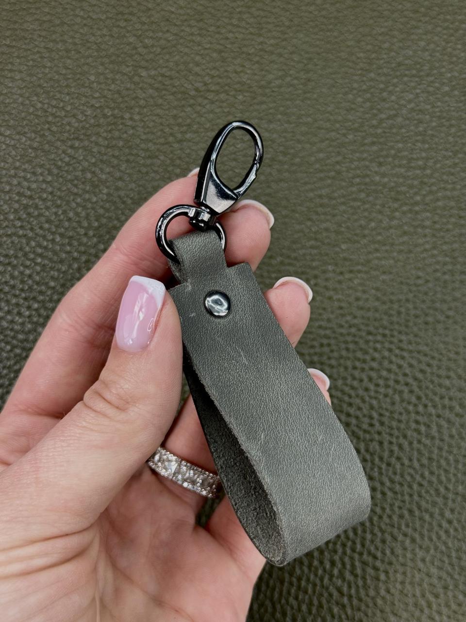 A hand with pink manicured nails holds a dark gray leather keychain with a black swivel clasp against a textured olive green background.