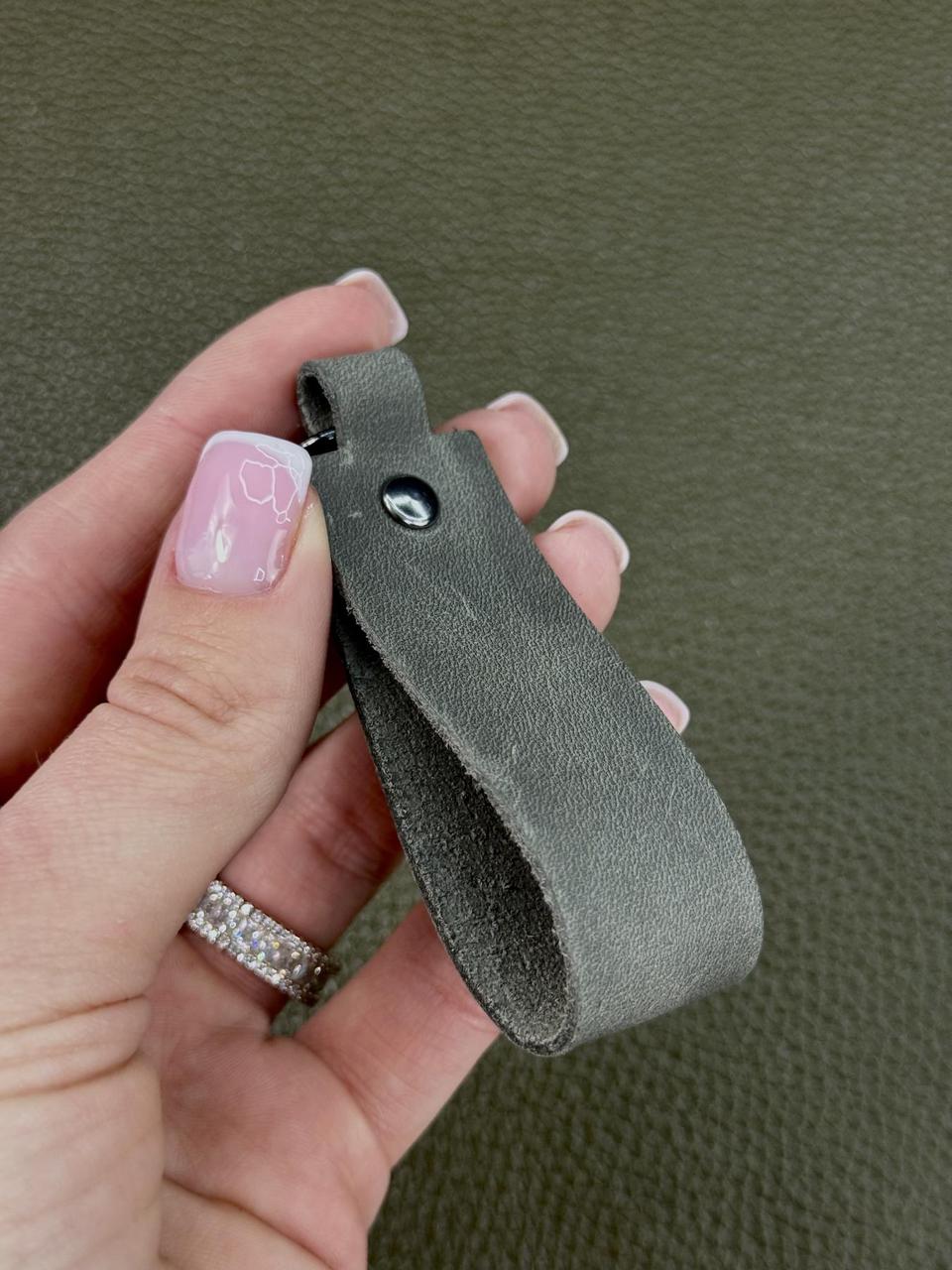 A close-up of a hand holding a small gray leather keychain or key ring against a textured olive green background.