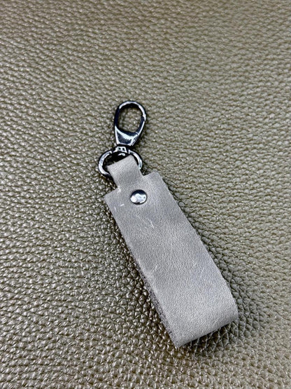 A silver-toned metal keychain or bag charm with a rectangular leather tag and a lobster clasp, placed on a textured beige surface.
