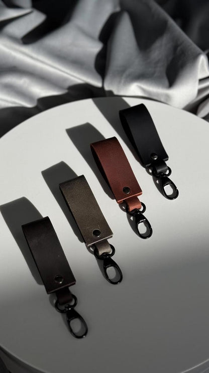 Three leather keychain clips in black, dark brown, and reddish-brown, arranged on a round white surface with draped gray fabric in the background.