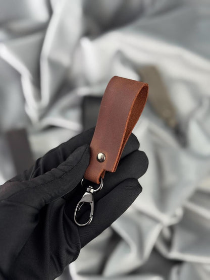 A close-up of a hand wearing a black glove holding a brown leather keychain strap with a silver metal clasp, set against a soft, draped gray fabric background.