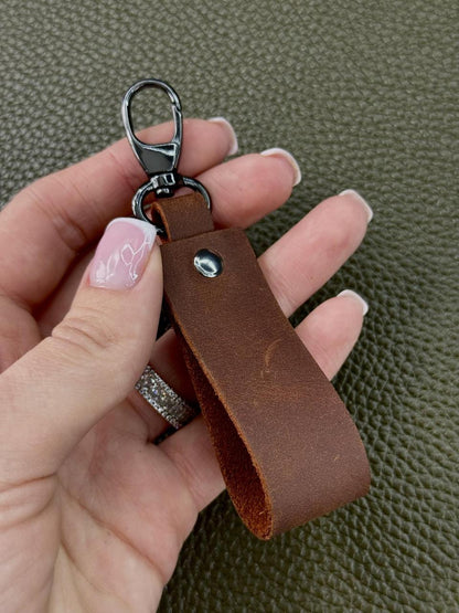 A close-up of a hand holding a brown leather keychain with a silver-toned metal clasp against a textured olive green background.