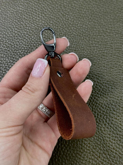 A close-up of a hand holding a brown leather keychain loop with a black metal carabiner clip, set against a textured olive green background.