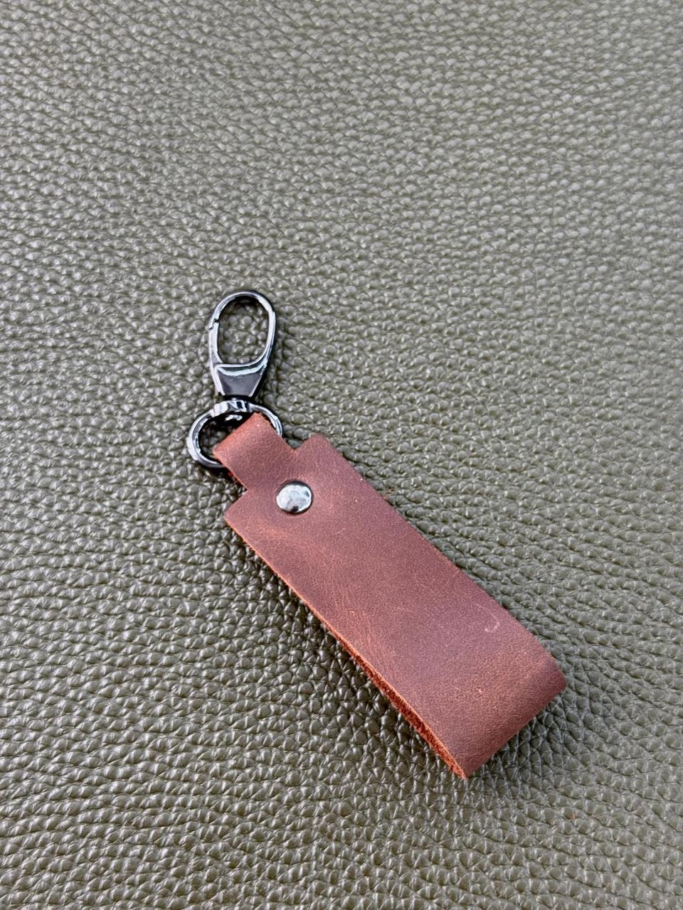 A brown leather keychain tag with a silver metal lobster clasp, shown on a textured olive green surface.