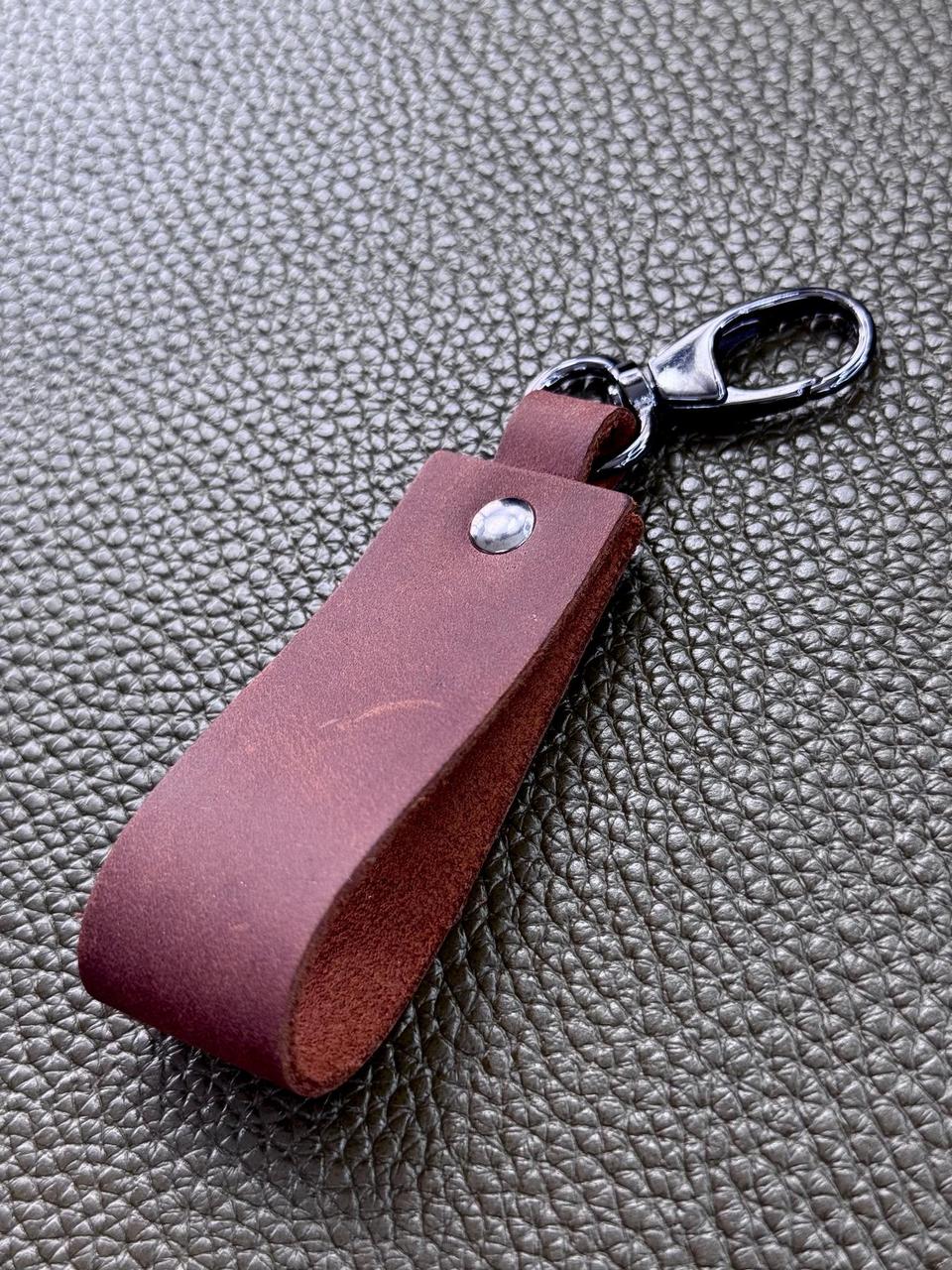 A dark reddish-brown leather keychain tag with a silver metal rivet and a silver lobster clasp, resting on a textured gray surface.
