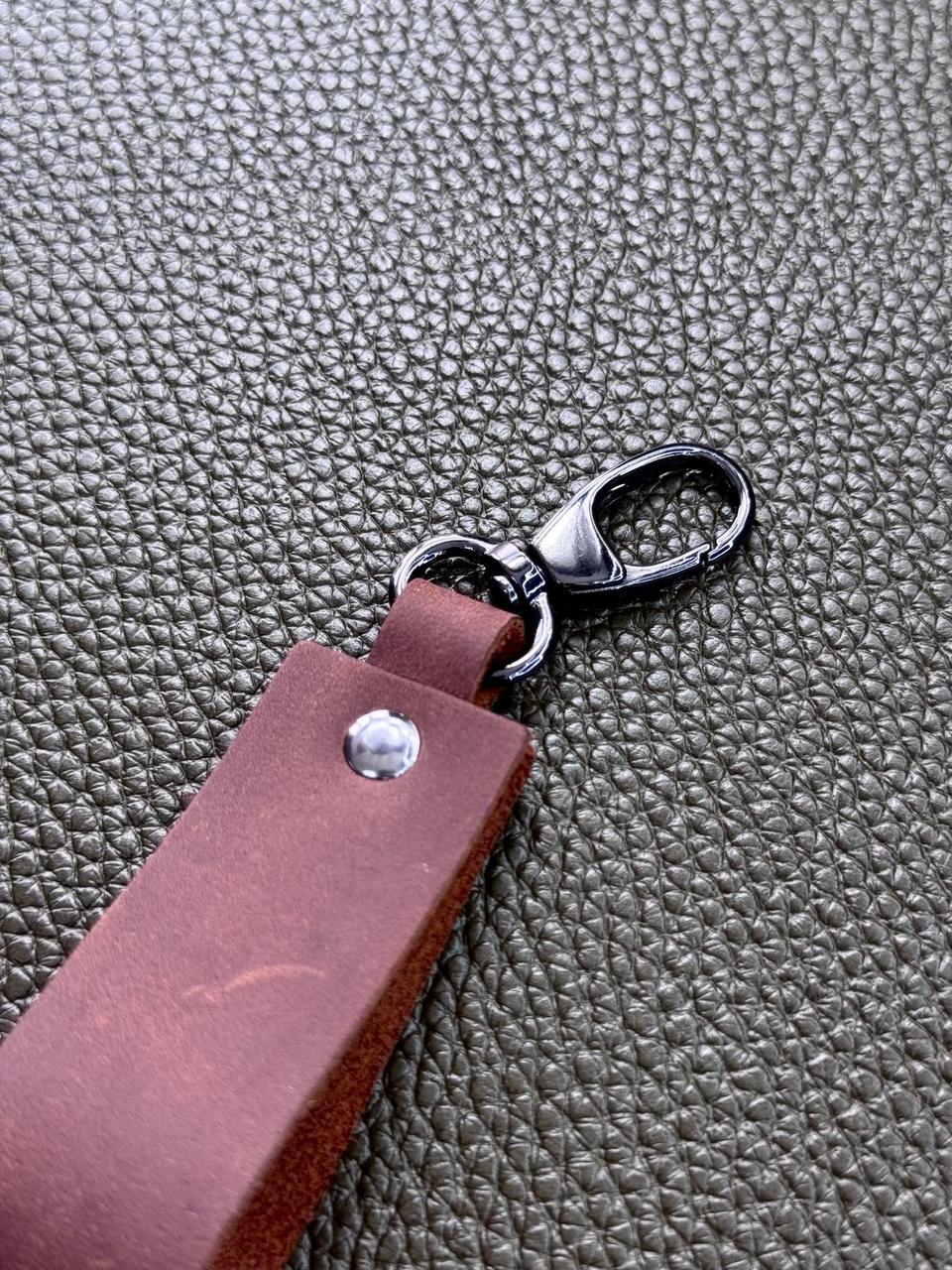A close-up of a brown leather keychain or bag strap with a silver metal lobster clasp, resting on a textured dark gray surface.