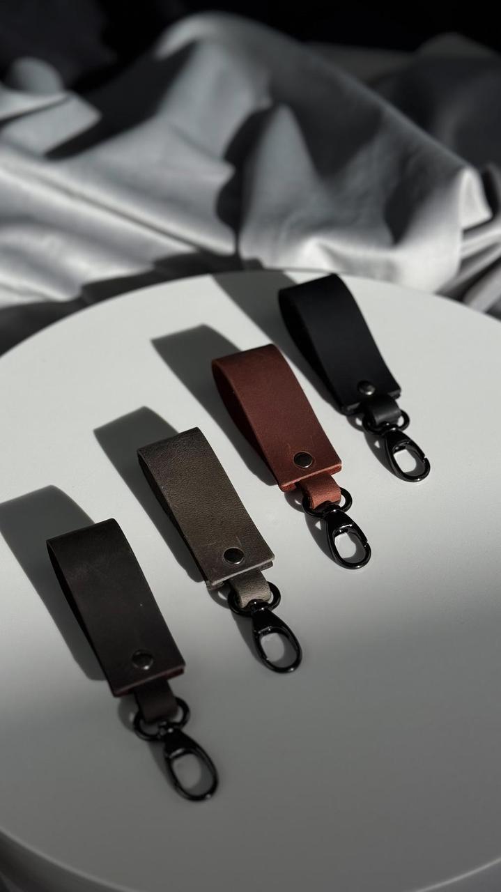 Four leather keychain pouches in black, brown, and dark gray, each with a metal lobster clasp, arranged on a white circular surface with draped gray fabric in the background.